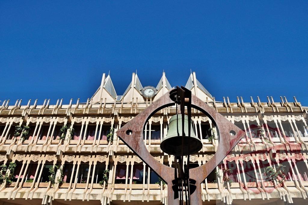 Modern bell sculpture in front of city hall architecture. Extremdura - Spain | Contemporary bell art integrating with detailed municipal building facade against blue sky - Realisiert mit Pictrs.com