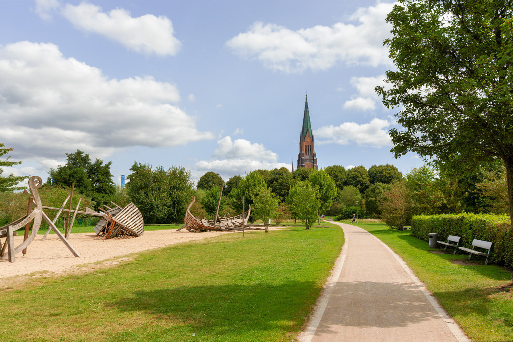 Wandbild: Weg über die Königswiesen in Schleswig  | Dieses Wandbild im Querformat zeigt einen Weg über die Königswiesen in Schleswig. Links neben dem Weg befindet sich ein schöner Spielplatz. In der Ferne ist der Schleswiger Dom zu sehen. Am blauen Himmel befinden sich einige sommerliche Wolken.  - Realisiert mit Pictrs.com