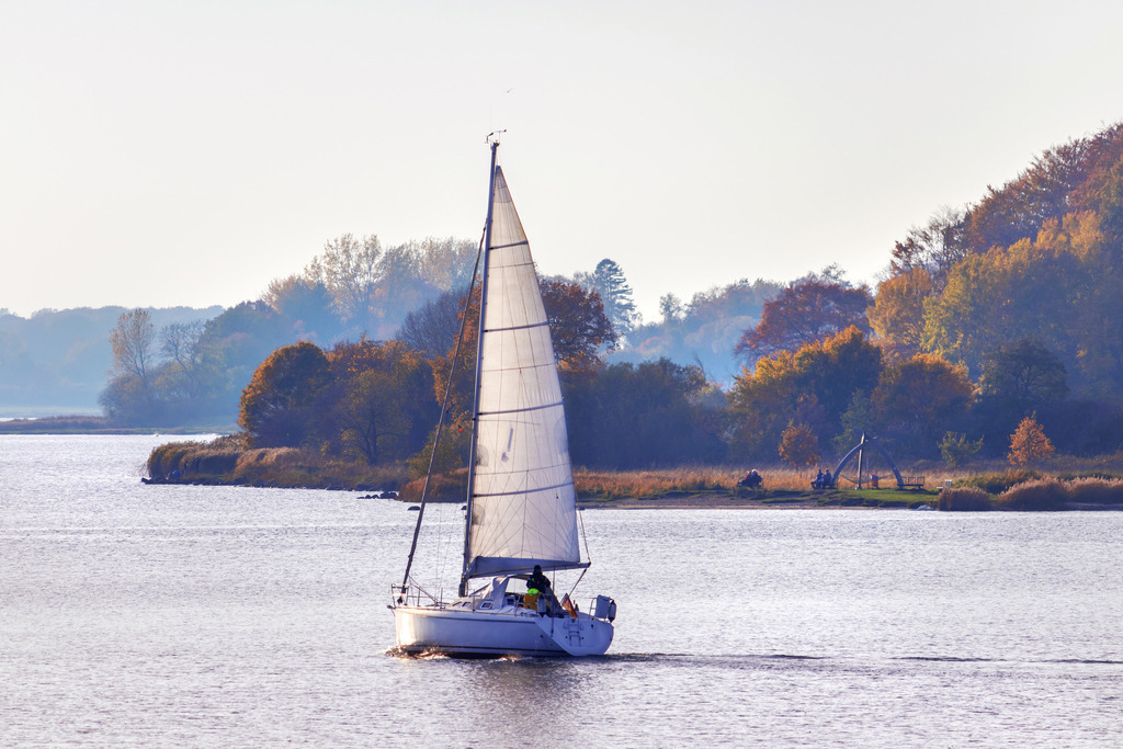 Wandbild: Segelboot auf der Schlei in Lindaunis | Dieses Wandbild im Querformat zeigt ein Segelboot auf der Schlei im Herbst. Auf der rechten Seite ist der Schleistrand Schneiderhaken zu sehen.  - Realisiert mit Pictrs.com