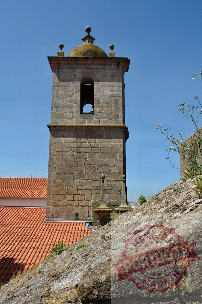 Stone bell tower standing tall under blue sky. Porto - Portugal | Old stone bell tower rising above terracotta roof with ancient wall in foreground - Realized with Pictrs.com
