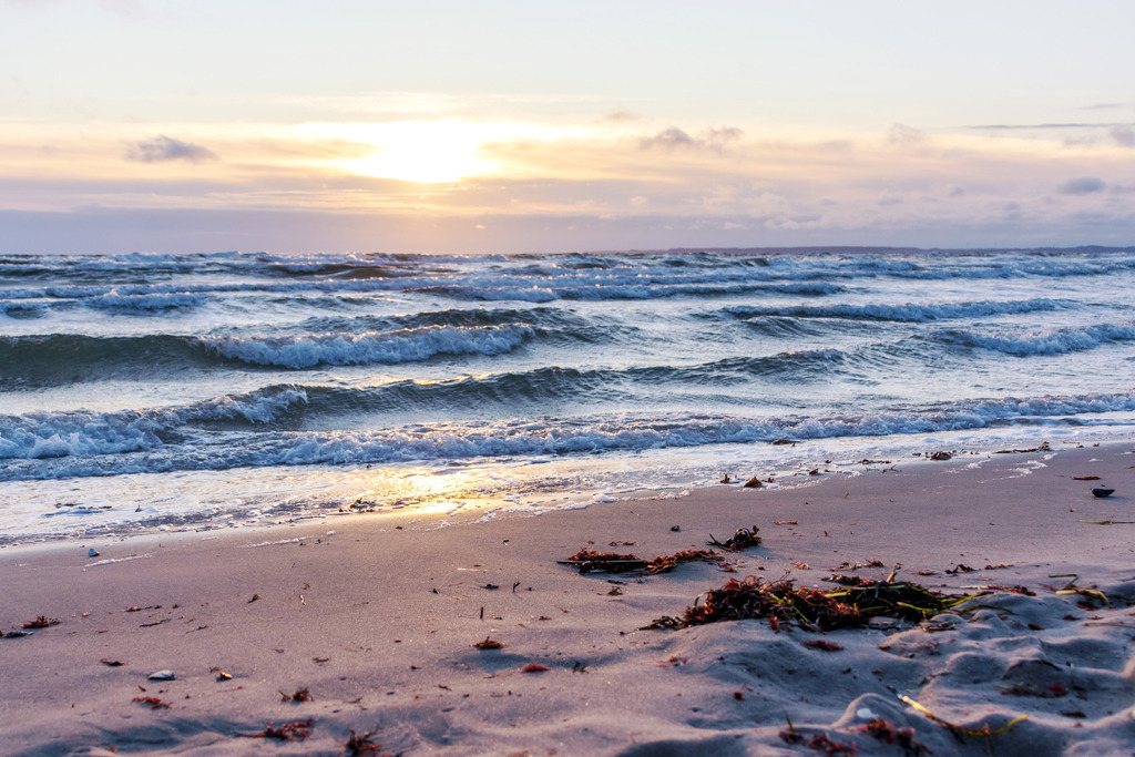 Wandbild: Sonnenaufgang an der Ostsee | Dieses Wandbild im Querformat zeigt einen schönen Sonnenaufgang an der Ostsee. Im Vordergrund ist der Sandstrand zu sehen, der teilweise von dem Licht der aufgehenden Sonne angeleuchtet wird. Auf dem Meer sind zahlreiche Wellen. Am Himmel scheint die Sonne durch Wolken am Horizont.  - Realisiert mit Pictrs.com