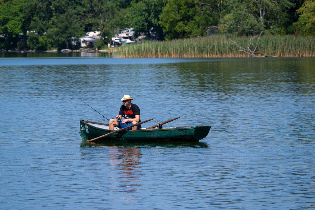 Sommerauszeit am Staffelsee: Naturidylle im Blauem Land | Ein Moment purer Entschleunigung vor der Kulisse von Murnau: Mitten auf dem sanft glitzernden Staffelsee sitzt ein Angler in seinem grünen Ruderboot und genießt die wohltuende Stille des Blauen Landes. Mit Strohhut, Sonnenbrille und Angelrute blickt er gelassen übers Wasser – fernab vom Hektik des Alltags. Im Hintergrund schmiegt sich das malerische, schilfbewachsene Ufer an die oberbayerische Voralpenlandschaft. Eine friedliche Sommeraufnahme, die Freiheit, Geduld und die zeitlose Seele des Staffelsees spürbar macht. - Realisiert mit Pictrs.com