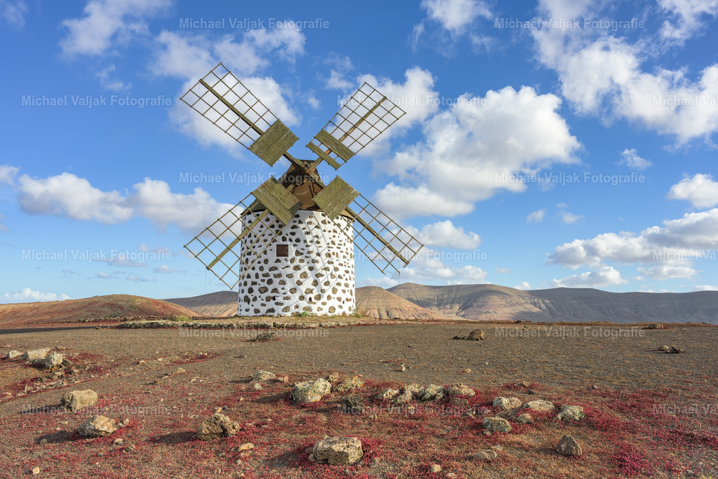 Windmühle in Villaverde auf Fuerteventura | In Villaverde steht die alte Windmühle ruhig über der weiten, hellen Landschaft des Nordens. Das Licht betont die klaren Linien des runden Mühlenkörpers und die hölzernen Flügel, während sich im Hintergrund die sanften Hügel der Vulkanlandschaft abzeichnen. Die Szene wirkt offen und weit, ein stiller Moment, in dem die Mühle wie ein fester Orientierungspunkt in der klaren Luft Fuerteventuras steht. - Realisiert mit Pictrs.com