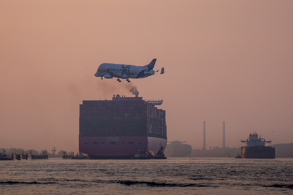 Begegnung der Giganten: Beluga XL & ONE Ingenuity | Der Airbus Beluga XL (Nummer 6) ist im Landeanflug auf das Werk in Finkenwerder, direkt über dem einlaufenden Containerschiff ONE Ingenuity. Das Bild zeigt das Zusammentreffen zweier markanter Transportmittel der modernen Logistik vor der Kulisse des Hamburger Hafens.  - Realisiert mit Pictrs.com