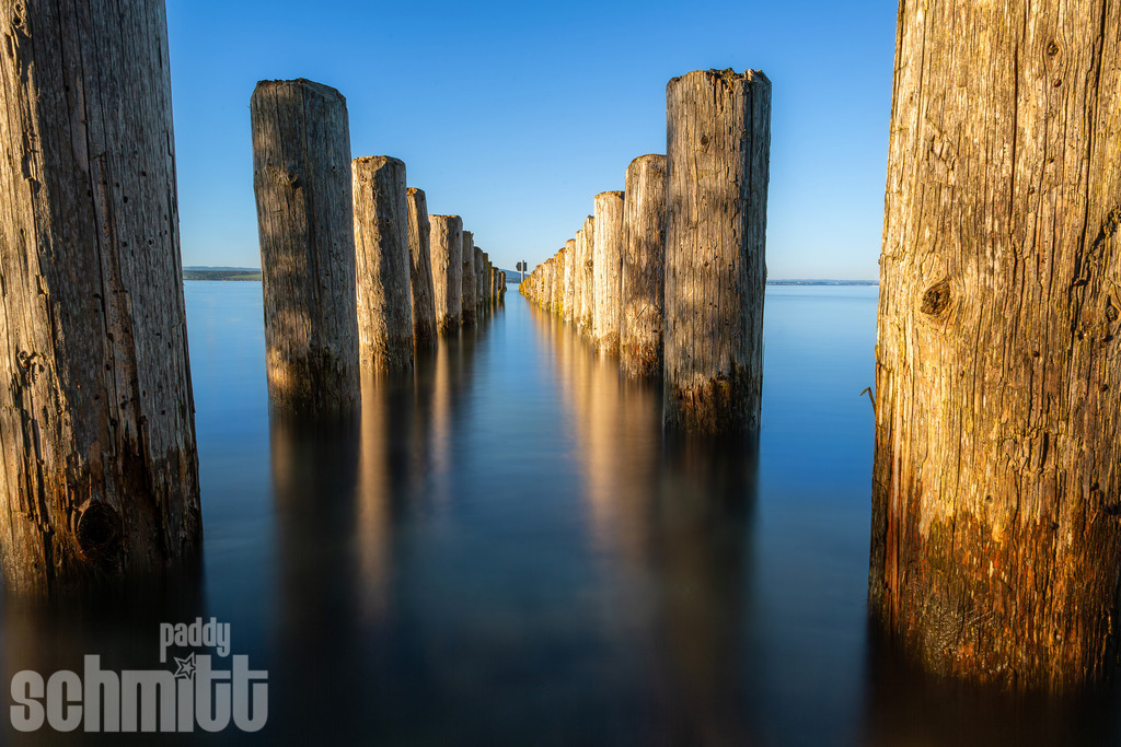 Holzpfosten am Bodenseeufer in der Schweiz | Holzpfosten am Schweizer Bodenseeufer stehen ruhig im Wasser und bildet einen natürlichen Blickfang. Die schlichte Szenerie mit dem weiten See vermittelt eine besondere Ruhe und die ursprüngliche Atmosphäre der Uferlandschaft. - Realisiert mit Pictrs.com