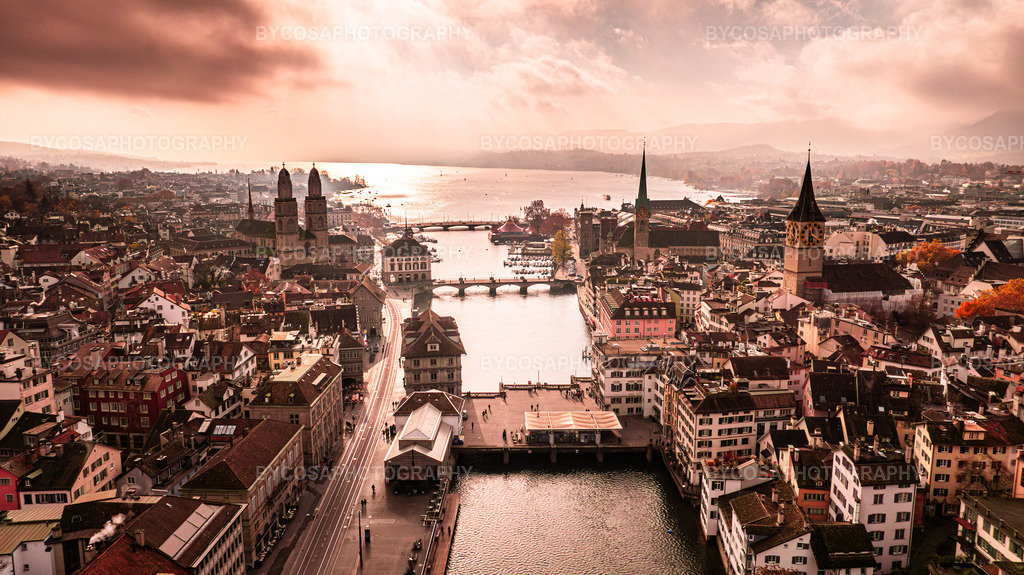 Golden Zürich _ Aerial Light Over the City | Ein atemberaubendes Luftpanorama von Zürich, eingefangen in einem seltenen Moment, in dem die Sonne durch die Wolken bricht und die gesamte Altstadt in warmes, goldenes Licht taucht. Das ikonische Trio – Grossmünster, Fraumünster und St. Peter – erstrahlt über den historischen Dächern, während der See zu einem glitzernden goldenen Horizont wird.Dieser FineArt-Druck verleiht jedem Raum Luxus, Wärme und pure Magie – die perfekte Wahl für moderne Interieurs, Büros und alle Zürich-Liebhaber. - Realisiert mit Pictrs.com
