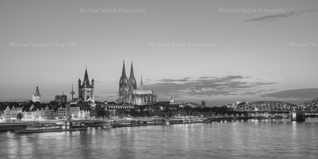 Die Skyline von Köln am Abend schwarz-weiß | Die Skyline von Köln, eingefangen in einem schwarz-weißen Bild am Abend, bietet einen zeitlosen Blick auf die Stadt. Die Konturen der Kölner Domspitzen heben sich markant gegen den Himmel ab, während die Lichter der Stadt ein sanftes Leuchten entlang des Rheins verbreiten. Diese Ansicht verbindet historische Architektur mit der Lebendigkeit der modernen Stadt und schafft ein visuelles Erlebnis, das sowohl nostalgisch als auch zukunftsweisend ist. - Realisiert mit Pictrs.com
