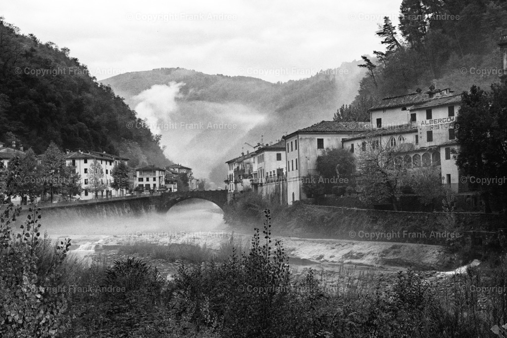 Bagni di Lucca Toskana | Schwarz weiß Fotografie von Bagni die Lucca in der Toskana an einem nebligen Tag. Blick über den Fluss Lima, über dem Nebel zieht, zu den Häusern der Altstadt. Im Hintergrund die Berge der Toskana die im Nebel verblassen. - Realisiert mit Pictrs.com