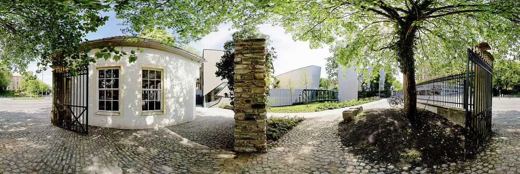Akzisehaus und Felix-Nussbaum-Haus, Osnabrück | Das historische Akzisehaus, ein Osnabrücker Zollhaus aus dem Jahr 1817, steht in direkter Nachbarschaft zum Felix-Nussbaum-Haus. Es gehört zum Museumsquartier Osnabrück und wird für kulturelle Veranstaltungen genutzt. In den Jahren 2026/27 dient es auch vorübergehend als Trauzimmer des Standesamtes. Das Bild vereint die architektonischen Gegensätze an diesem Ort: Das alte traditionelle Zollhaus vor dem im Hintergrund sichtbaren modernen asymmetrischen Museumsbau. Auch die Baumaterialien kontrastieren stark: Warme Naturmaterialien im Vordergund (Kopfsteinpflaster, Sandsteinpfeiler, Putzfassade) gegenüber Beton, Glas und Zinkblech beim Felix-Nussbaum-Haus. Die Spannung der Gegensätze löst sich unter einem lichtdurchfluteten Blätterdach in einer heiteren und friedlichen Atmosphäre auf. Für einen Moment gehört der Ort ganz allein den Betrachtenden und den eigenen Gedanken. Das Bild entstand am 08. Mai 2011, ein Gedenktag, an dem im Jahr 1945 der Zweite Weltkrieg in Europa endete. - Realisiert mit Pictrs.com