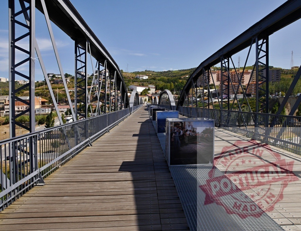 Metal truss bridge displaying outdoor art exhibition | Pedestrian bridge with a wooden deck showing an outdoor art exhibition on a sunny day - Realisiert mit Pictrs.com