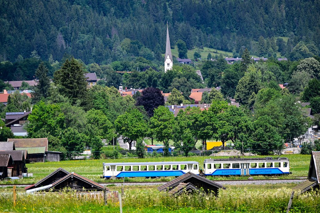 Unterwegs zum höchsten Gipfel: Ein Zug voller Träume | Ein Bild, das die Sehnsucht nach Abenteuer weckt und das Herz erwärmt: Inmitten einer saftig grünen, blühenden Sommerwiese zieht die blau-weiße Bayerische Zugspitzbahn gemächlich ihre Bahnen. Im Hintergrund schmiegt sich der idyllische Ort mit seinen roten Dächern in die Landschaft, überragt vom eleganten, spitzen Kirchturm, der wie ein Wegweiser in den Himmel ragt. Dichte Nadelwälder säumen die Hänge und strahlen eine tiefe Ruhe aus. Diese Aufnahme fängt die perfekte Harmonie aus traditioneller Nostalgie, Naturidylle und der Vorfreude auf die Bergwelt ein – eine Reise, die den Alltag hinter sich lässt. - Realisiert mit Pictrs.com