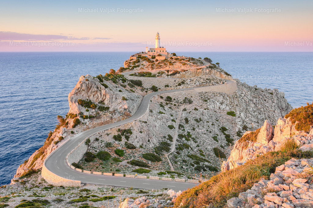 Far de Cap Formentor auf Mallorca | Im Abendlicht zeigt sich der Far de Cap Formentor in einer stillen, fast zurückhaltenden Atmosphäre. Die Sonne sinkt langsam Richtung Horizont und legt einen warmen Schimmer auf die felsige Spitze, sodass die Formen der Klippen weich, aber dennoch klar erkennbar bleiben. Der Leuchtturm steht wie ein heller Punkt über dem Meer, das in sanften Tönen schimmert und nur leichte Bewegungen zeigt. - Realisiert mit Pictrs.com