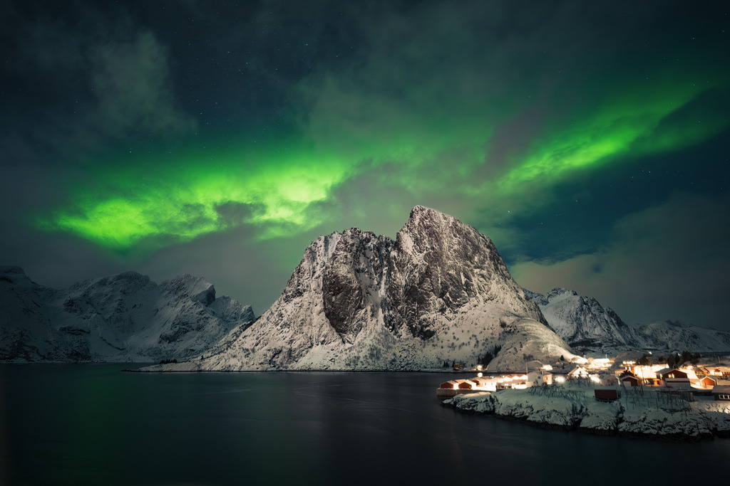 Polarlicht über den Bergen Nordnorwegens | Grünes Polarlicht zieht über einen markanten Berg und die Lichter eines kleinen norwegischen Küstenortes. Die winterliche Nachtlandschaft verbindet arktische Natur und warme Siedlungslichter. - Realisiert mit Pictrs.com