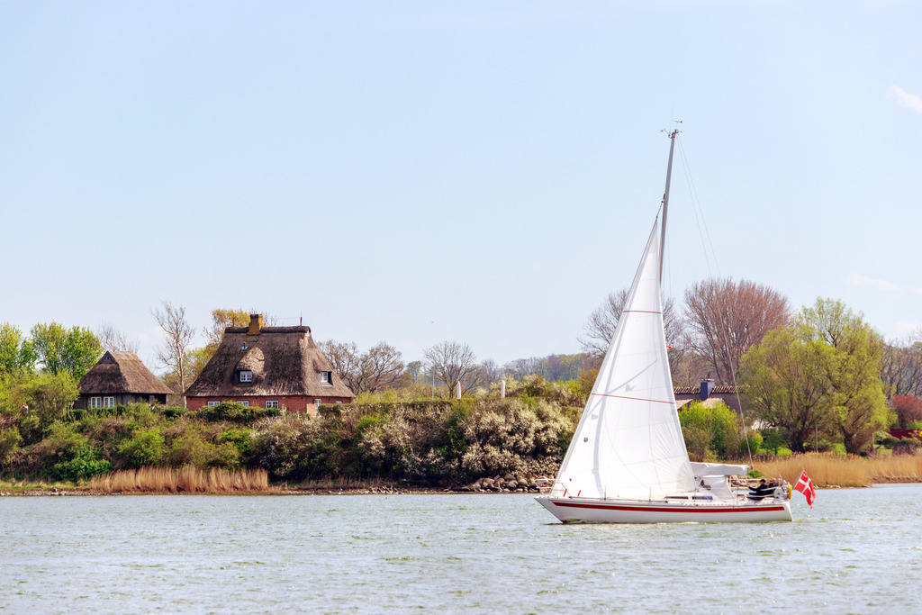 Wandbild: Segelboot auf der Schlei in Arnis | Dieses Wandbild im Querformat zeigt ein Segelboot auf der Schlei in Arnis im Frühling. Direkt am Ufer der Schlei steht ein malerisches Haus mit Reetdach. Der blaue Himmel ist wolkenlos. - Realisiert mit Pictrs.com